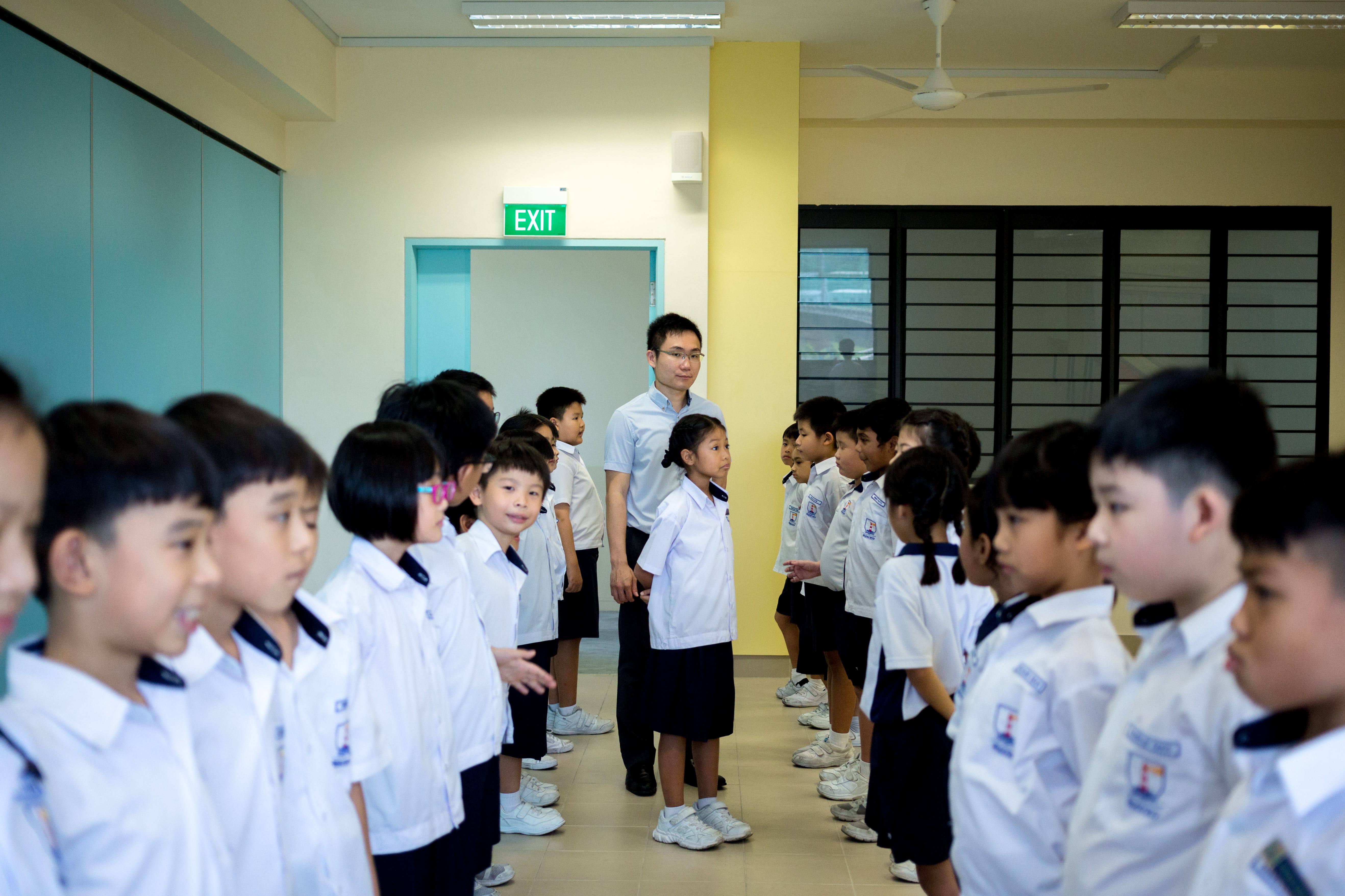 Teacher stands between two lines of schoolchildren in uniforms in a hallway under an EXIT sign.